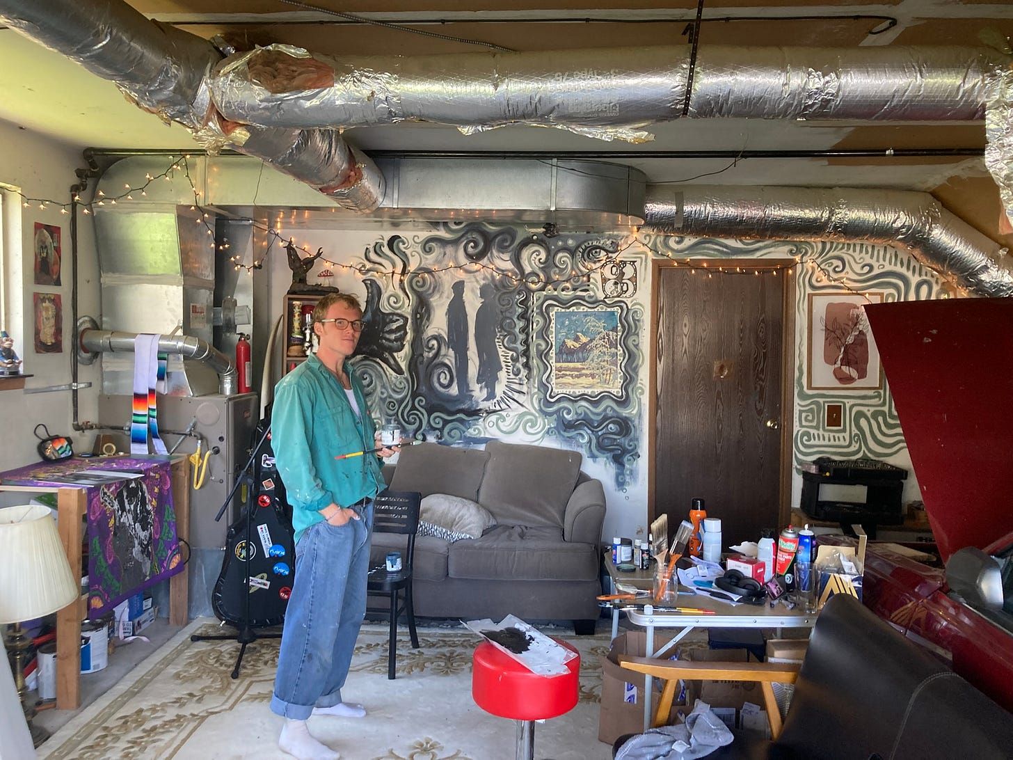A man mid-mural in a garage, holding a brush and a small tin, in a paint-spattered teal jacket and socks. Behind him the wall is covered in black and white swirls with two silhouetted figures standing in them, painted around a small framed landscape. Foil-wrapped ductwork crosses the ceiling, string lights are strung along the wall, and a folding table of paint cans and spray bottles stands beside a grey couch and a red stool.