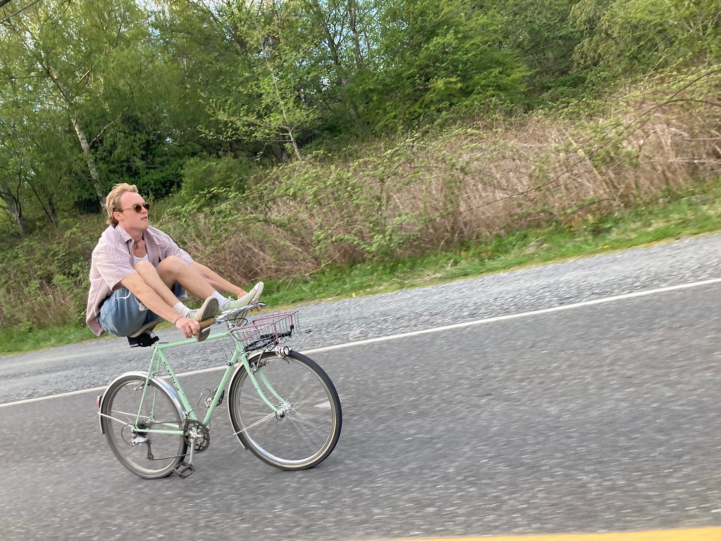 A man coasting a pale green bike down a road with both feet up on the handlebars, sunglasses on, open checked shirt, hands off the bars. The asphalt and the brush along the shoulder are blurred with the speed.
