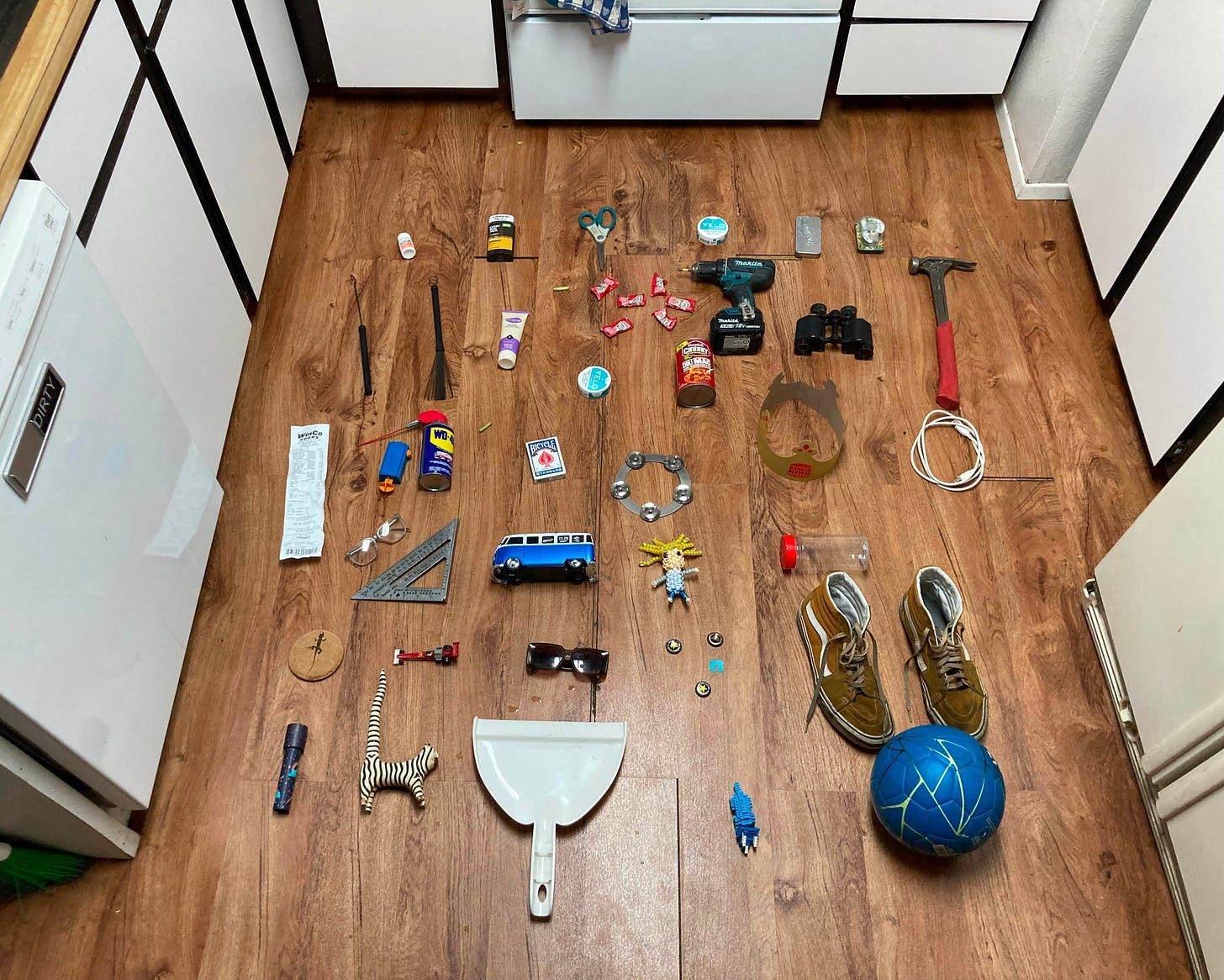 Objects laid out in loose rows on a wood floor between a dishwasher and the kitchen cabinets, shot from above. Among them a cordless drill, a claw hammer, binoculars, scissors, a can of WD-40, a deck of Bicycle cards, a tambourine jingle, a speed square, reading glasses, sunglasses, a blue toy VW bus, a beaded figure, a paper crown, a coiled white cable, a tin of cherry gummies, loose sweets, a zebra figurine, a dustpan, brown Vans and a blue football.