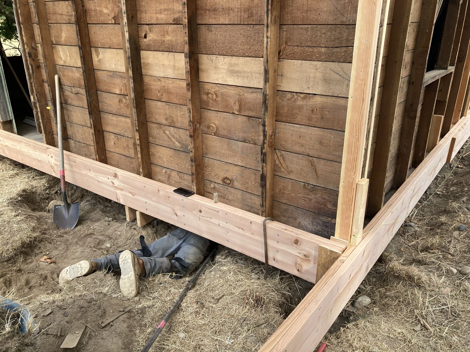 The corner of the house with its siding off, bare board sheathing and studs showing. New pale timber has been bolted along the base as a lifting beam. A pair of boots and legs in grey trousers stick out from underneath, the rest of the man in under the house. A shovel stands in the dirt, a phone sits on the beam.