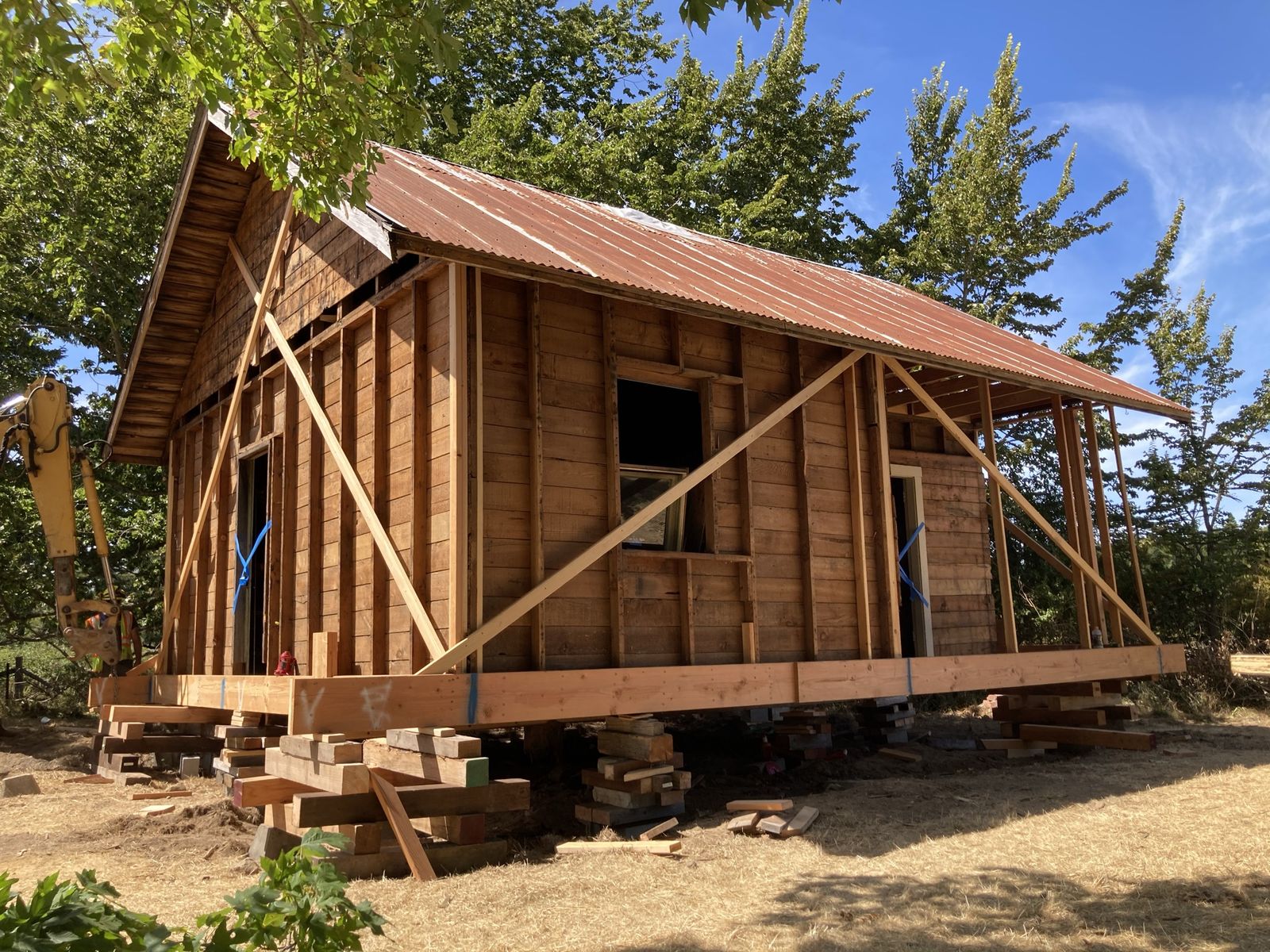 The house lifted clear of the ground on stacks of cribbing timber, seen from the corner. Rusted corrugated roofing, bare board walls with new diagonal braces nailed across them, window openings empty. A yellow excavator arm at the left, dry grass, blue sky.