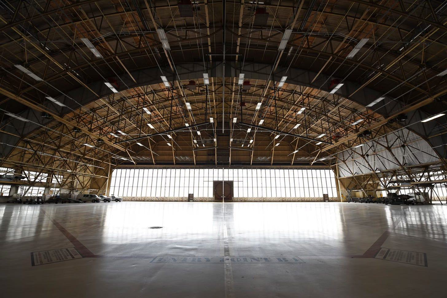 The inside of an empty airplane hangar. An arched roof of wooden and steel lattice trusses runs the length of it, hung with rows of fluorescent lights. A wall of tall windows fills the far end with white daylight. The concrete floor is polished enough to reflect it, with ENTRY POINT B stencilled across the middle, and vehicles and aircraft sit in shadow along both side walls.